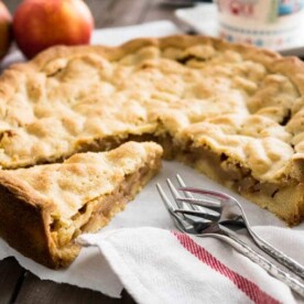 A covered German Apple Cake on a white dishtowel with a slice cut out but left in place with some forks. There's a mug and 2 apples in the background.