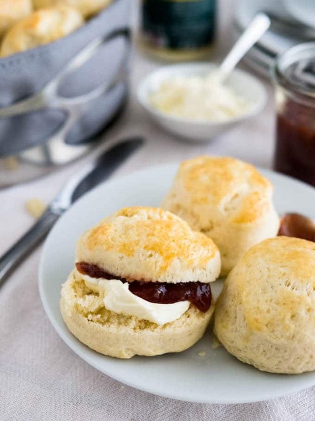 A white plate with English scones and a spoon of marmalade. One of the scones is halved and has clotted cream and strawberry jam on it. There\'s a small white bowl of clotted cream with a spoon and a jar of strawberry jam in the background.