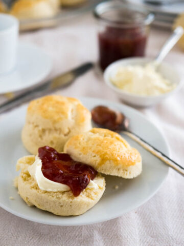 A white plate with English scones and a spoon of marmelade. One of the scones is halved and has clotted cream and strawberry jam on it. There's a small white bowl of clotted cream with a spoon and a jar of strawberry jam in the background.