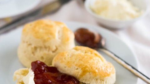 A white plate with English scones and a spoon of marmelade. One of the scones is halved and has clotted cream and strawberry jam on it. There's a small white bowl of clotted cream with a spoon and a jar of strawberry jam in the background.