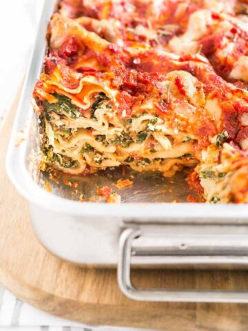 A metal baking pan with spinach lasagna on a wooden cutting board. There's a piece of it missing.