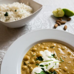 A white plate of chickpea curry with sour cream and cilantro on a silver tablecloth. There's a square bowl of rice, some almonds and two squeezed halves of lime next to it.
