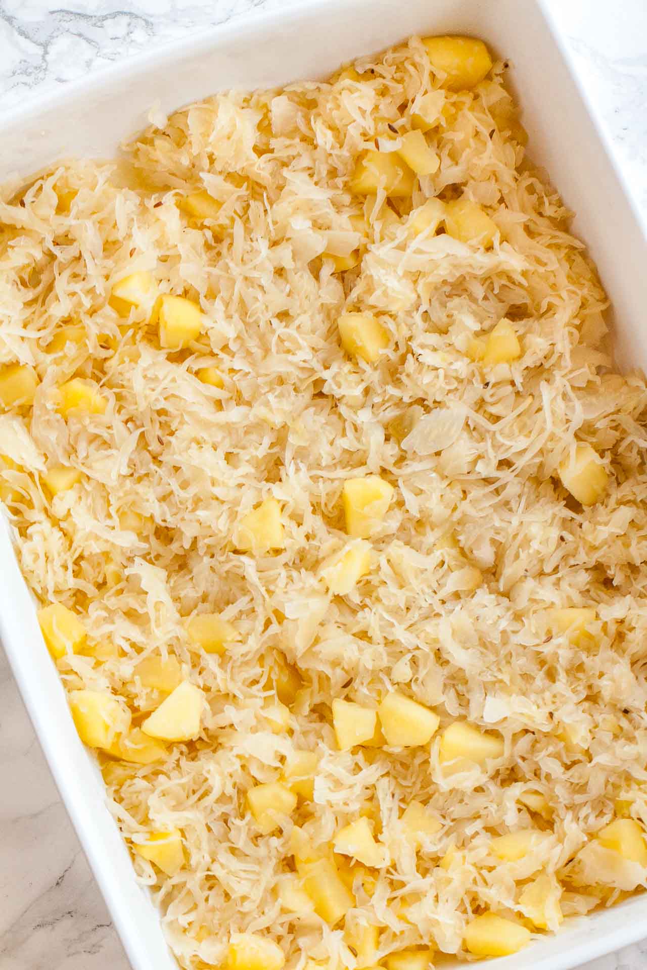 A layer of apple pieces and sauerkraut in a white baking dish on a marble surface.