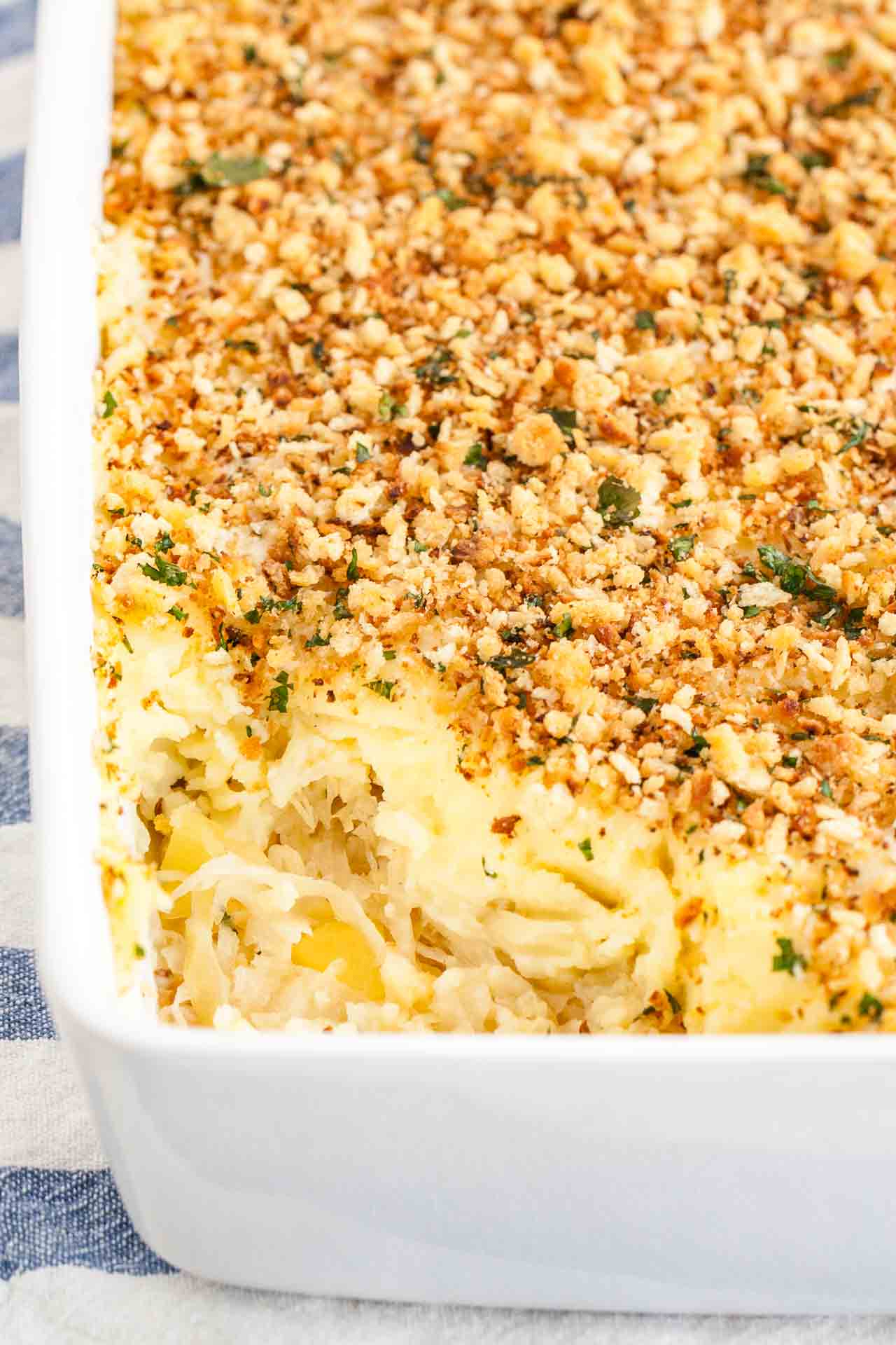 Close-up of a white baking dish with sauerkraut mashed potato casserole on a white and blue dishtowel. A portion of the casserole has been removed.