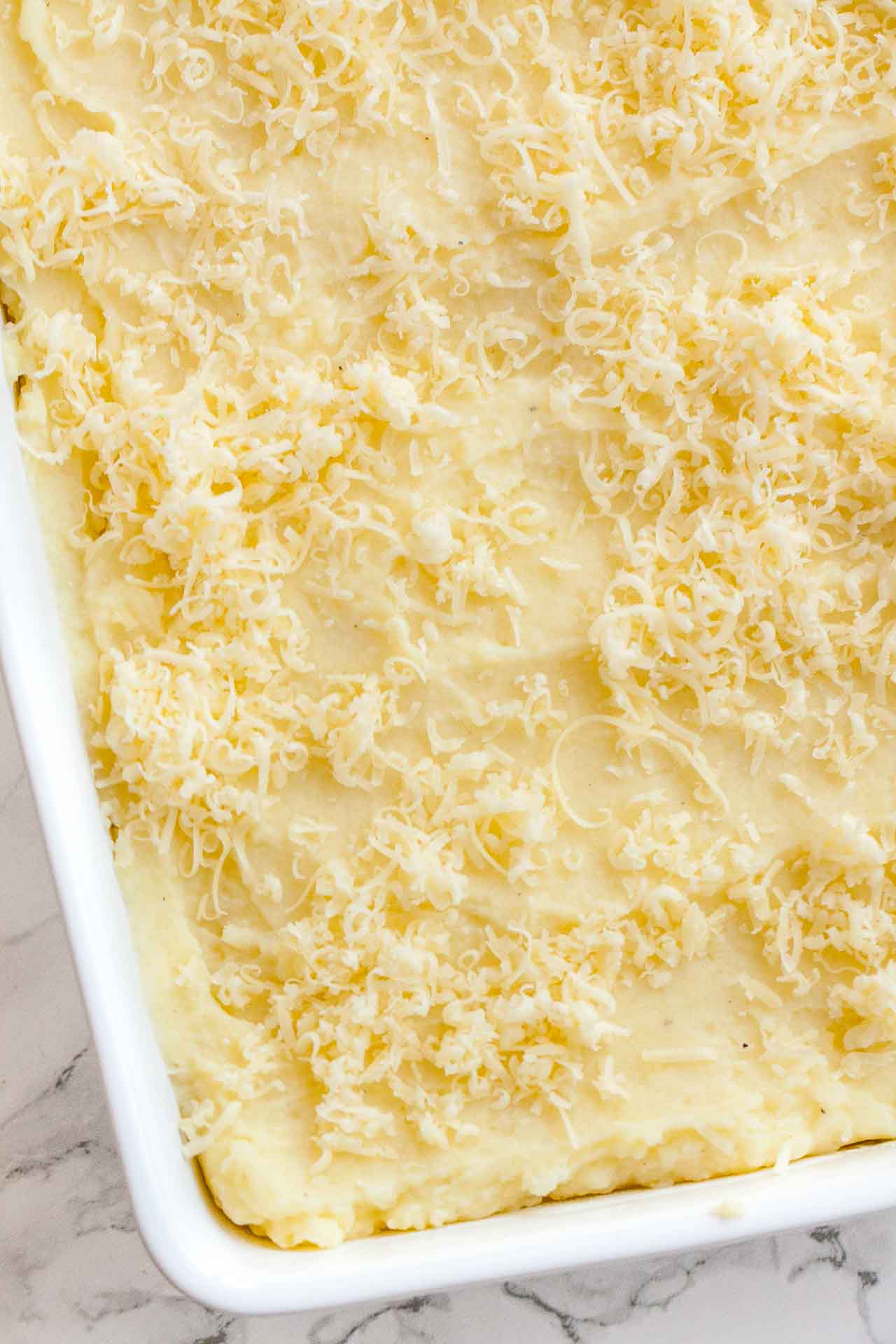 A layer of mashed potatoes topped with cheese in a white baking dish on a marble surface.