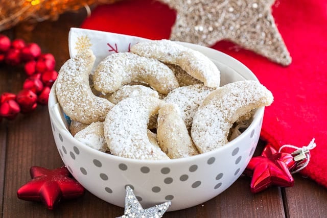 A white bowl with silver dots with a napkin, of Vanillekipferl cookies. Next to it, there are several Christmas stars.