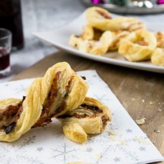 A festive napkin with a bacon and cheese puff pastry twists on a wooden cutting board. There's a white, rectangular plate in the background with more of the puff pastry twists.