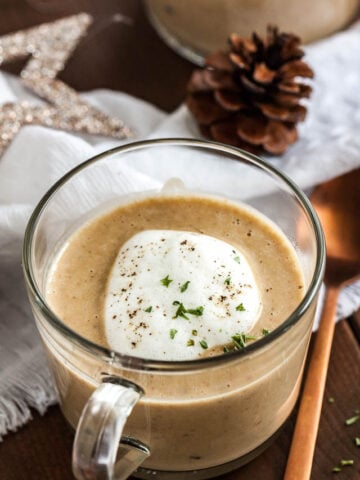 Close-up of a glass cup of chestnut soup with truffle froth on a wooden table with a bronze spoon next to it. There's a white tablecloth with Christmas ornaments in the background.