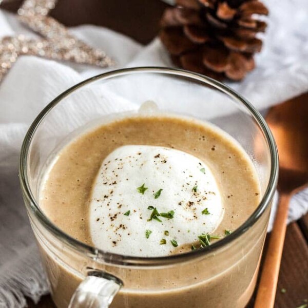 Close-up of a glass cup of chestnut soup with truffle froth on a wooden table with a bronze spoon next to it. There's a white tablecloth with Christmas ornaments in the background.