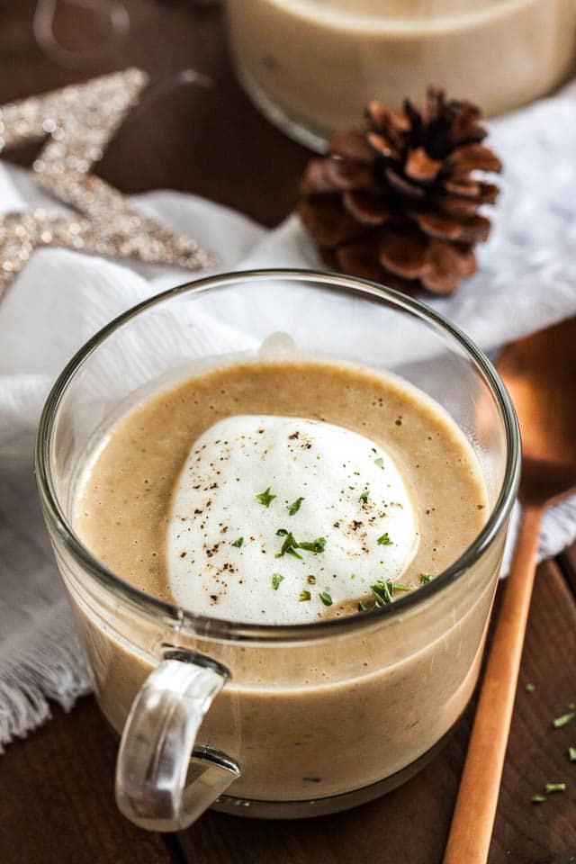 Close-up of a glass cup of chestnut soup with truffle froth on a wooden table with a bronze spoon next to it. There\'s a white tablecloth with Christmas ornaments in the background.
