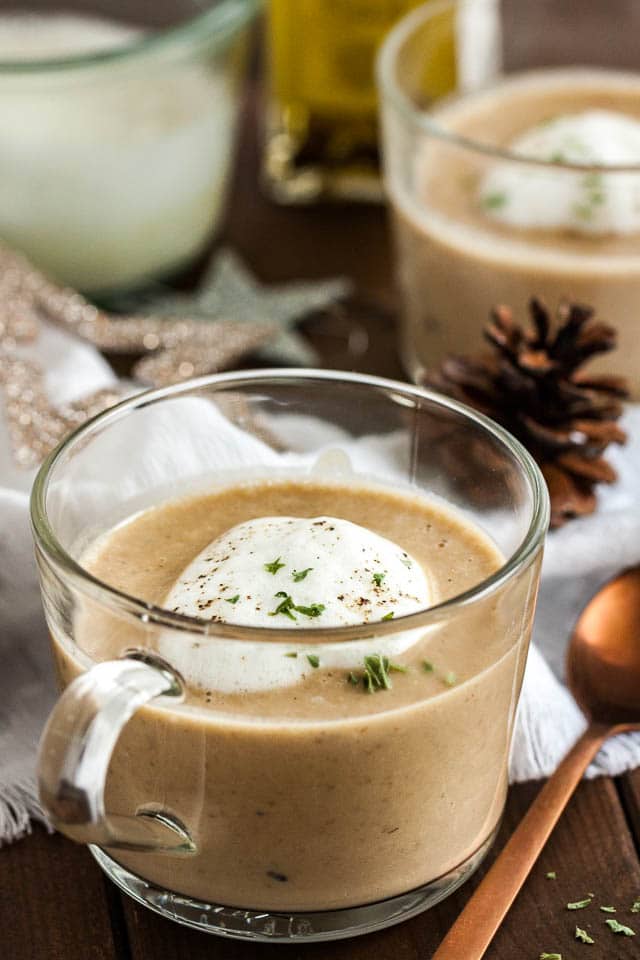 A glass cup of chestnut soup with truffle froth on a wooden table with a bronze spoon next to it. There\'s a white tablecloth with Christmas ornaments and another cup of soup in the background.
