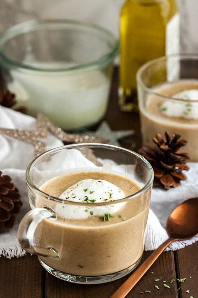 A glass cup of chestnut soup with truffle froth on a wooden table with a bronze spoon next to it. There\'s a white tablecloth with Christmas ornaments and another cup of soup in the background.