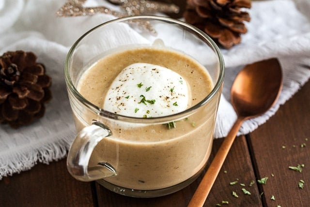 A glass cup of chestnut soup with truffle froth on a wooden table with a bronze spoon next to it. There\'s a white tablecloth with Christmas ornaments in the background.