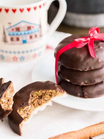 a stack of three chocolate-covered lebkuchen with a red bow on a white plate next to a lebkuchen broken in half. There's a cup in the background