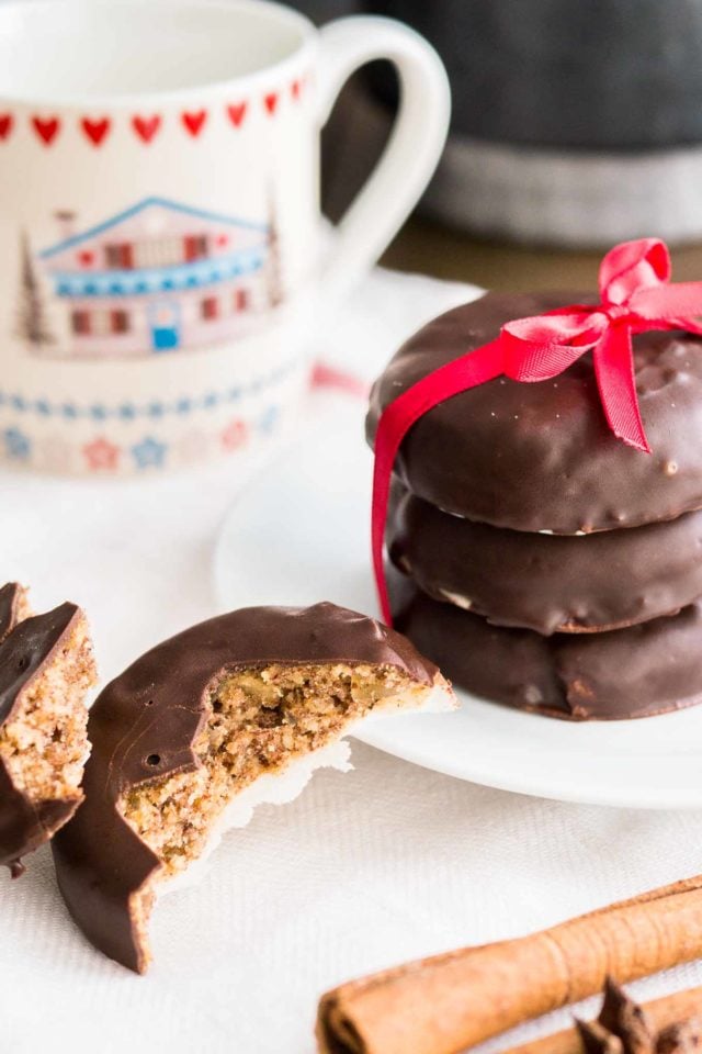 a stack of three chocolate-covered lebkuchen with a red bow on a white plate next to a lebkuchen broken in half. There\'s a cup in the background