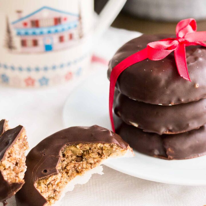 a stack of three chocolate-covered lebkuchen with a red bow on a white plate next to a lebkuchen broken in half. There's a cup in the background