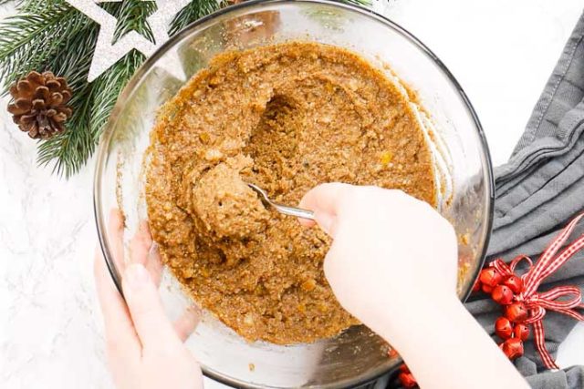 A glass bowl with lebkuchen dough next to some red jingle bells and some pine branches. A hand is scooping out dough with a teaspoon.