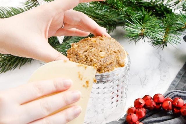 a hand is holding a lebkuchen. The wafer is on an upside-down whiskey tumbler and the dough is shaped into a shallow dome with a dough card. There are red jingle bells and pine branches in the background.