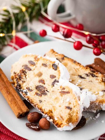 Two slices of christstollen on a white plate garnished with cinnamon sticks, cranberries and hazelnuts.