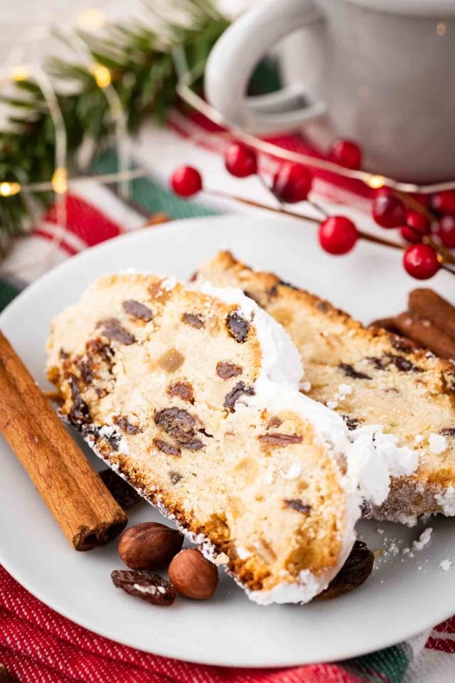 Two slices of christstollen on a white plate garnished with cinnamon sticks, cranberries and hazelnuts.