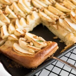 A baking sheet with apple cake with cinnamon butter topped with pine nuts on a black cooling rack. A cake server is lifting out a piece of the cake.