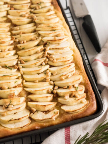 A baking sheet with apple cake with cinnamon butter topped with pine nuts on a black cooling rack. There's a knife and a white dishtowel with a red stripe next to it.