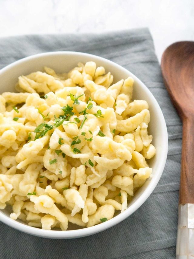 A white bowl of spaetzle garnished with parsley on a grey dishtowel next to a wooden spoon.