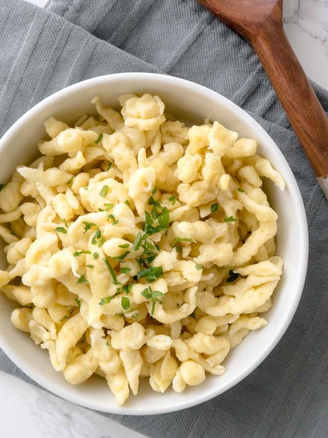 Top-down shot of a white bowl of spaetzle garnished with parsley on a grey dishtowel next to a wooden spoon.