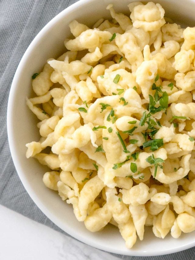 Close-up of a white bowl of spaetzle garnished with parsley on a grey dishtowel.