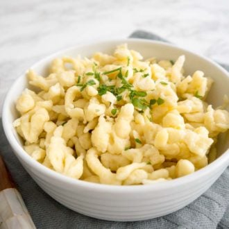 A white bowl of spaetzle garnished with parsley on a grey dishtowel next to a wooden spoon.