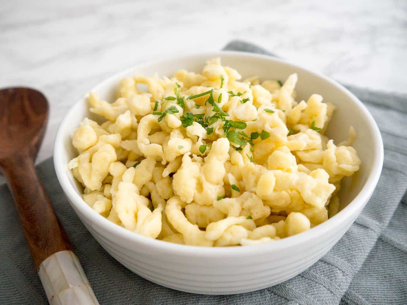 A white bowl of spaetzle garnished with parsley on a grey dishtowel next to a wooden spoon.