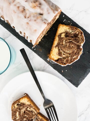 Top-down shot of a marble orange pound cake on a grey slab of granite with a slice of ti lying in front of it. There's a glass of milk and a white plate with another slice and a black fork next to it.