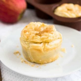 Close-up of a mini apple pie with dulce de leche on a white plate on a white dishtowel. In the background, there's a brown muffin pan with more of the pies and an apple.