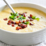 A white bowl of potato soup topped with bacon and parsley with a spoon in it on a white tablecloth.