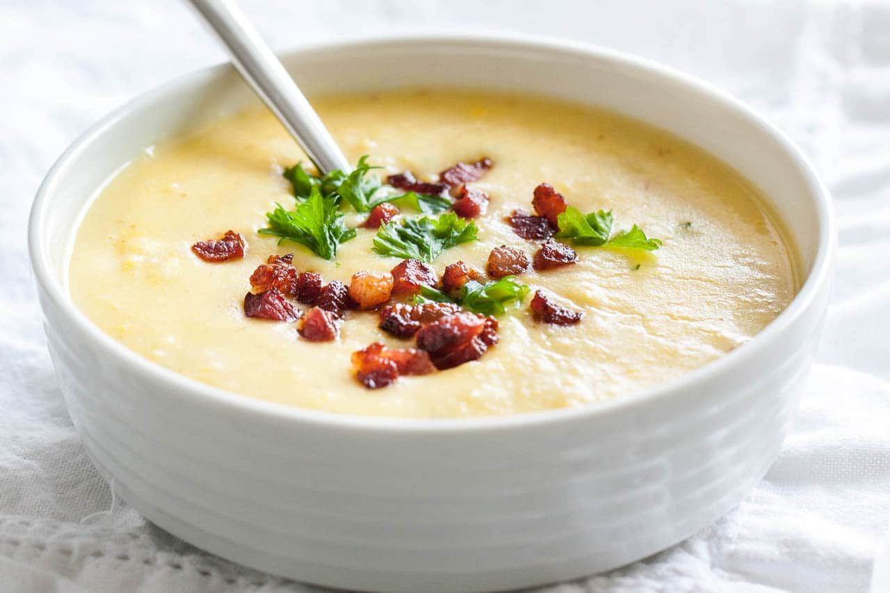 A white bowl of potato soup topped with bacon and parsley with a spoon in it on a white tablecloth.