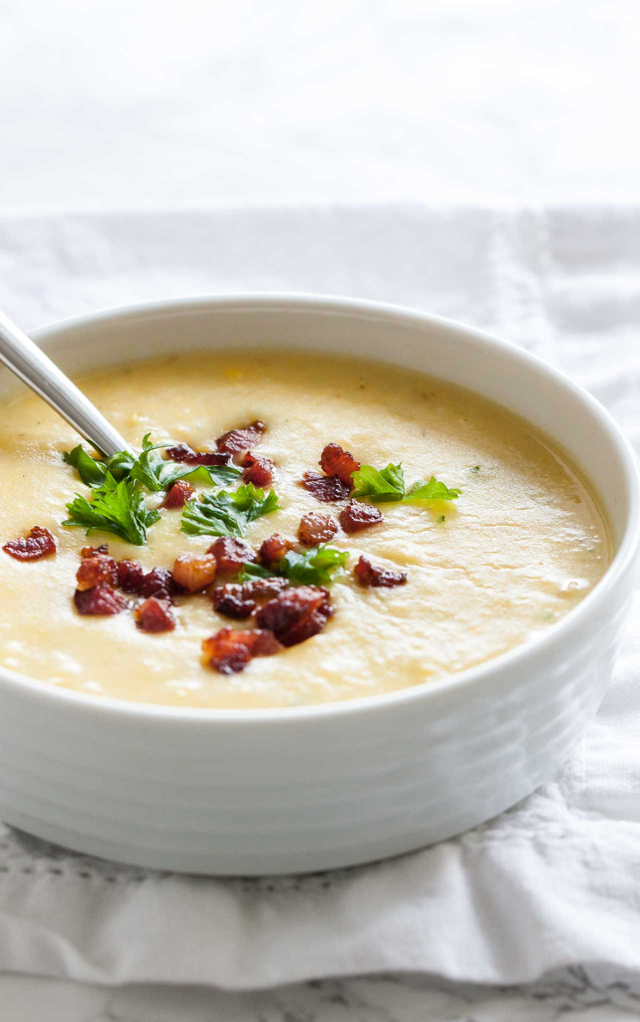 A white bowl of potato soup topped with bacon and parsley with a spoon in it on a white tablecloth.