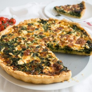 A spinach quiche with cheese and pine nuts on a springform bottom with a slice missing. There's a white plate with a slice in the background.