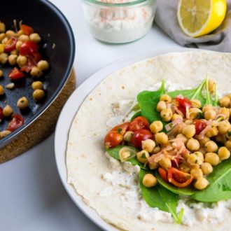 A white plate with spinach wrap with feta and chickpeas on it next to a pan with tomato chickpea mix. There's a jar with feta spread and half a lemon in the background.