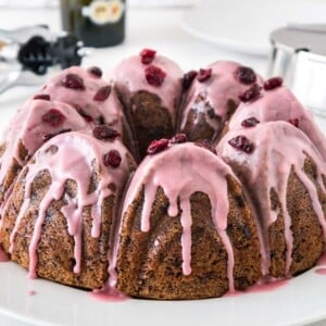 A chocolate cranberry bundt cake with pink frosting, garnished with dried cranberries on a white plate. In the background, there's a stainless steel sugar bowl and a wine opener.