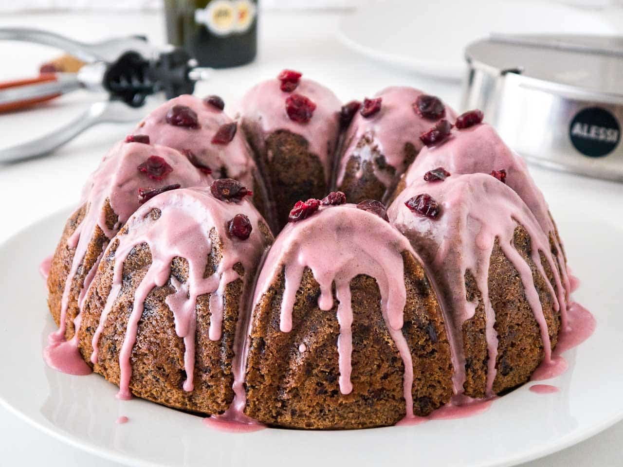 A chocolate cranberry bundt cake with pink frosting, garnished with dried cranberries on a white plate. In the background, there\'s a stainless steel sugar bowl and a wine opener.