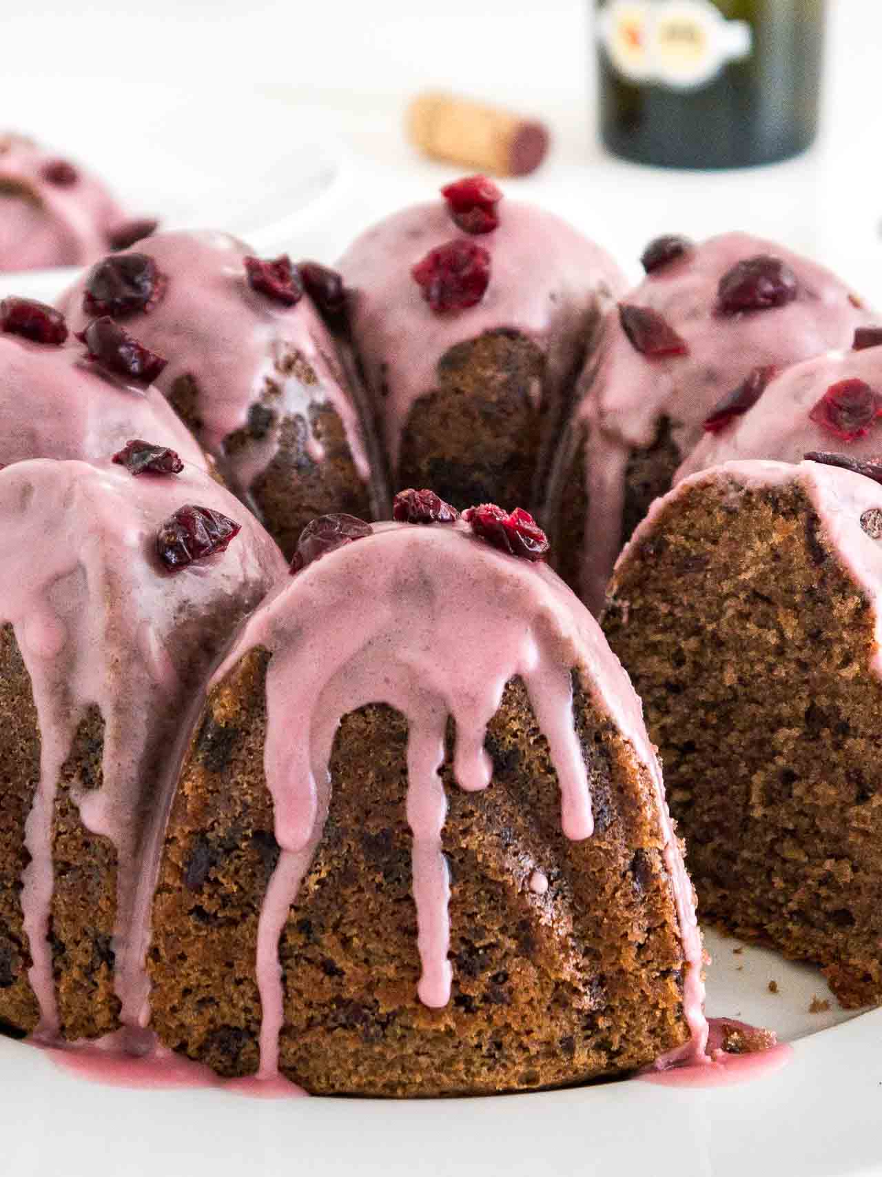 Close-up of a chocolate cranberry bundt cake with pink frosting, garnished with dried cranberries on a white plate. A piece of the cake has been cut out.