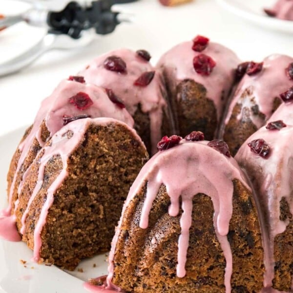 A chocolate cranberry bundt cake with pink frosting, garnished with dried cranberries on a white plate. A piece of the cake has been cut out. In the background, there's a stainless steel sugar bowl and a wine opener.
