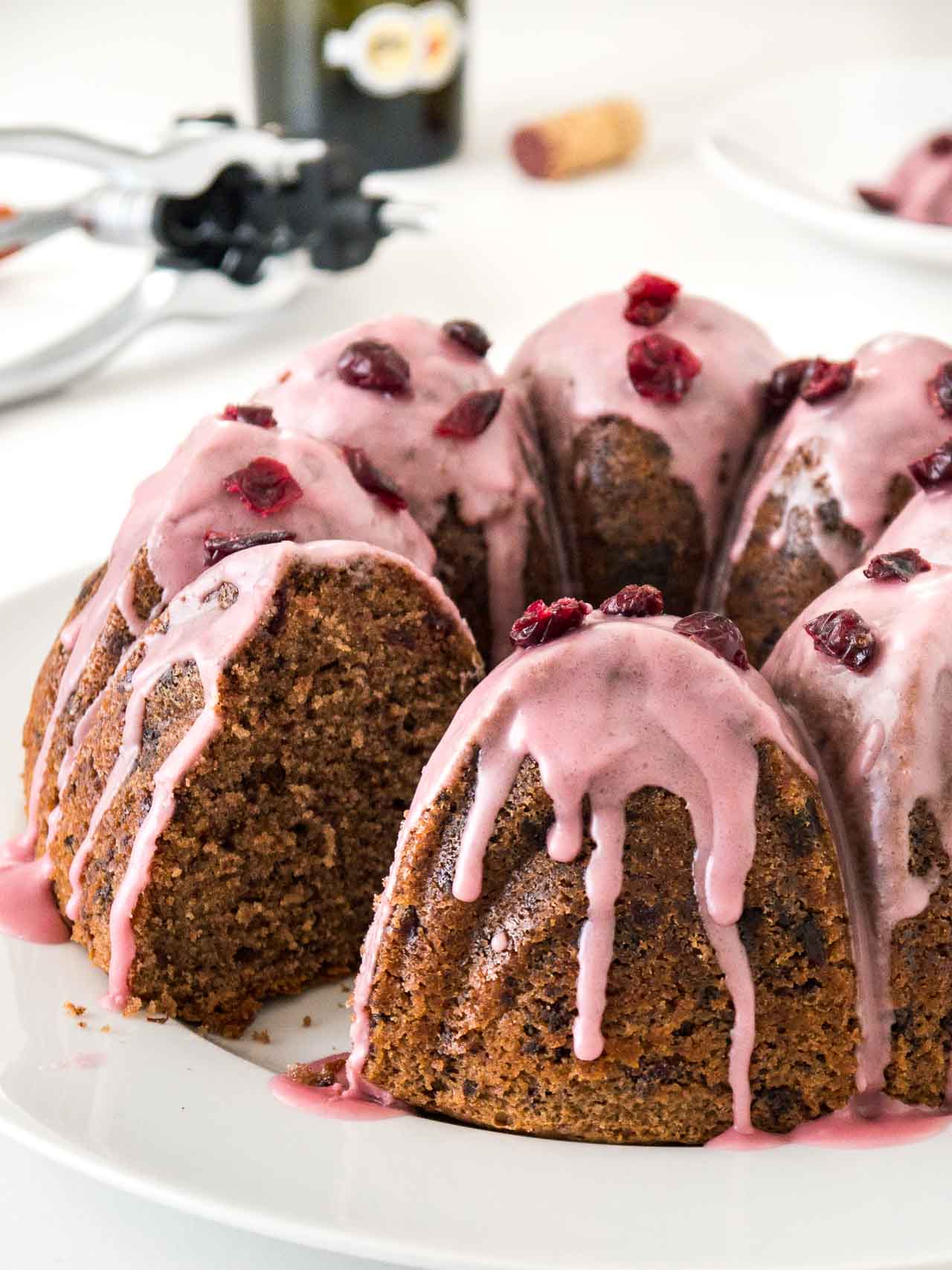 A chocolate cranberry bundt cake with pink frosting, garnished with dried cranberries on a white plate. A piece of the cake has been cut out. In the background, there\'s a stainless steel sugar bowl and a wine opener.