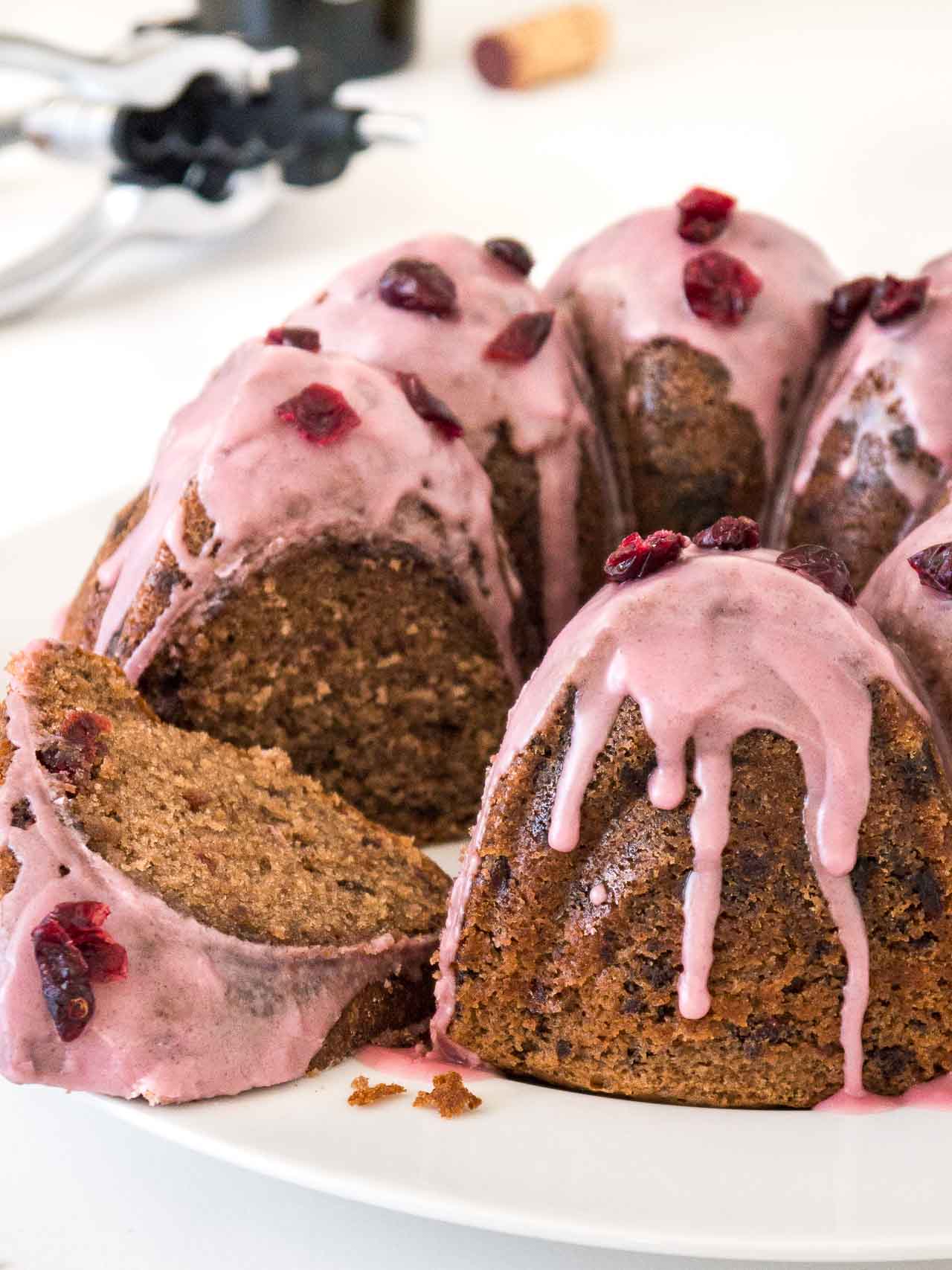A chocolate cranberry bundt cake with pink frosting, garnished with dried cranberries on a white plate. A piece of the cake has been cut out and is lying in front.