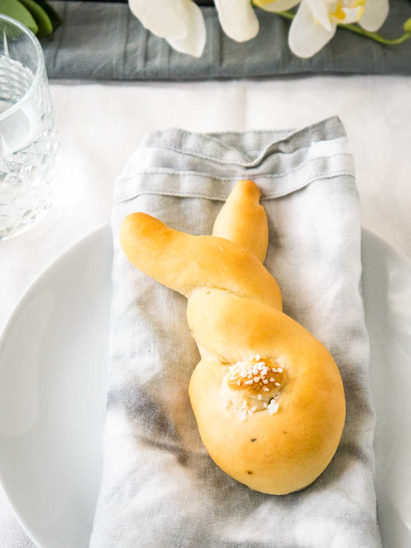 Bread shaped like a bunny, with a cottontail from dough on a grey dishtowel on a white plate next to a glass of water.