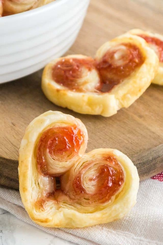 Close-up of a heart-shaped puff pastry palmier leaning against a wood cutting board with more palmiers in the background.