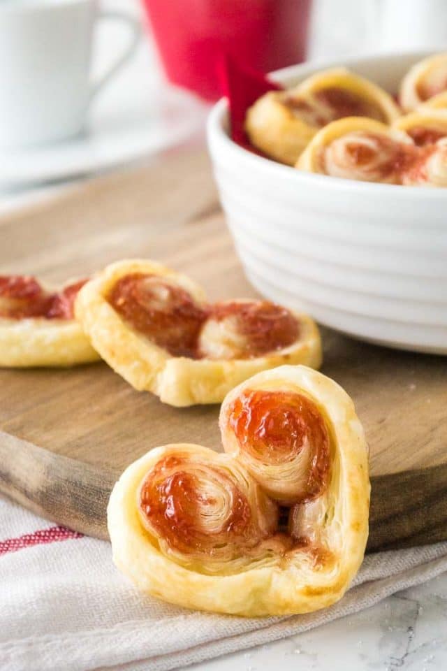 Close-up of Heart-shaped puff pastry palmiers. One is in the front leaning against a wooden cutting board, others are lying on the cutting board and in a white bowl on the board.