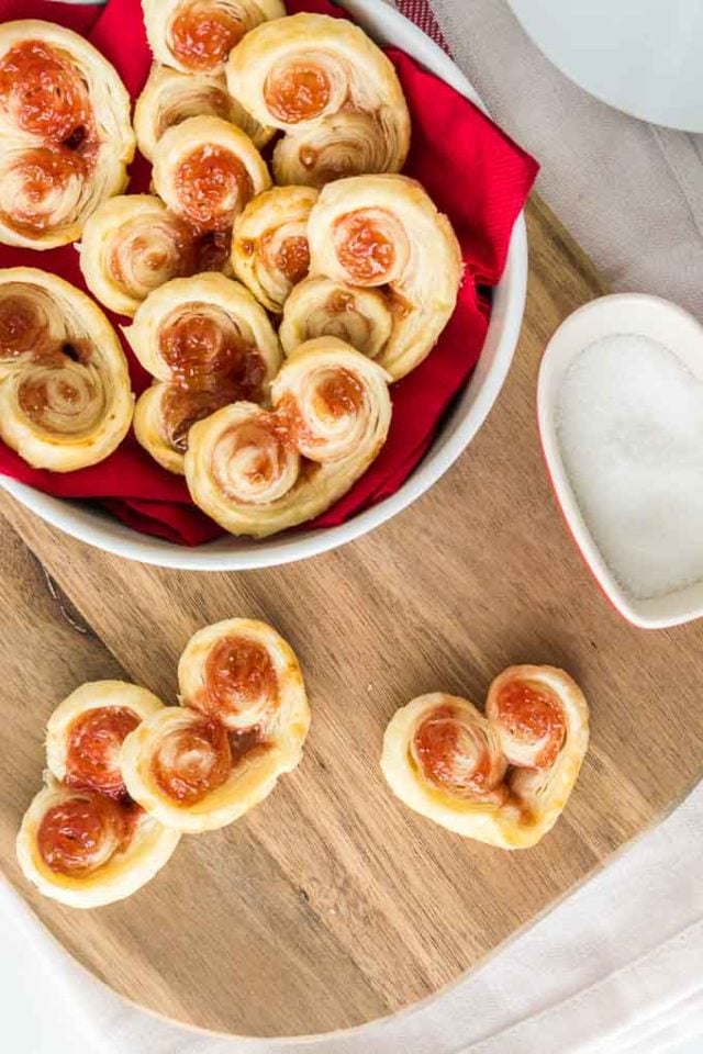 Top-down shot of heart-shaped puff pastry palmiers. Three are on a cutting board and the rest are in a white bowl lined with a red napkin.