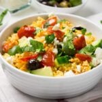 Greek couscous salad with cucumber, tomato, olives, basil and feta in a white bowl on a white dishtowel. There is a white bowl of olives and a white bowl with basil in the background.