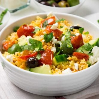 Greek couscous salad with cucumber, tomato, olives, basil and feta in a white bowl on a white dishtowel. There is a white bowl of olives and a white bowl with basil in the background.